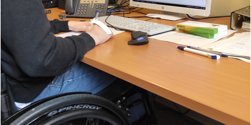 ein junger Mann sitzt im Rollstuhl im Büro an einem Schreibtisch 