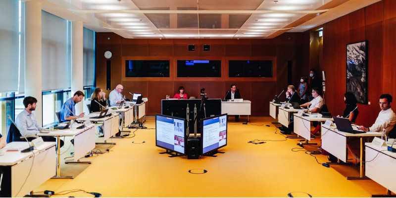 10 Personen sitzen in einem Saal an im Kreis angeordneten Tischen mit Laptops und Mikrofonen, in der Mitte große Bildschirme