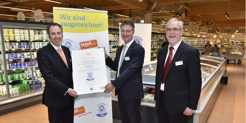 drei Männer in Anzügen stehen nebeneinander in einem Supermarkt und lächeln in die Kamera, zwei halten gemeinsam eine Urkunde mit dem "Blauen Engel" darauf , im Hintergrund ein Plakat mit dem "Blauen Engel" und der Aufschrift "Wir sind ausgezeichnet!"