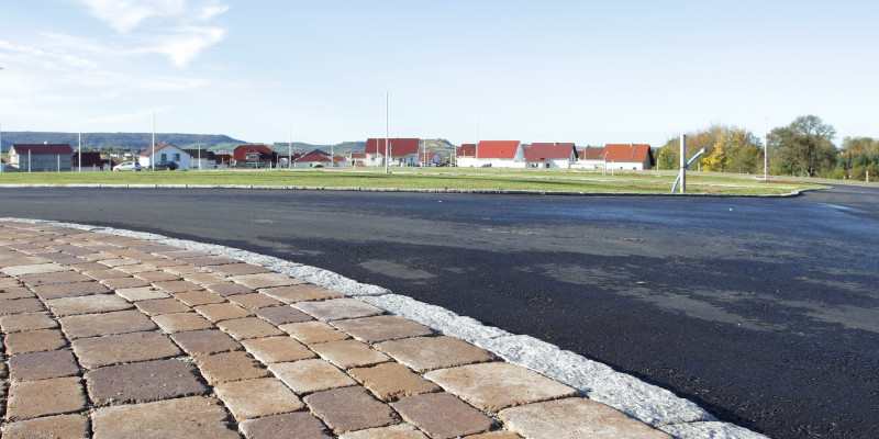 asphaltierte Straße und gepflasterter Weg, im Hintergrund ein Neubaugebiet "auf der grünen Wiese"