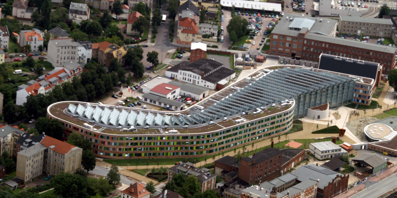 Das schlangenförmige UBA-Dienstgebäude in Dessau-Roßlau von oben fotografiert, zu sehen ist die Fassade aus Holz und bunten Glasscheiben und die schräg aufgestellten Solarzellen auf dem gesamten Flachdach