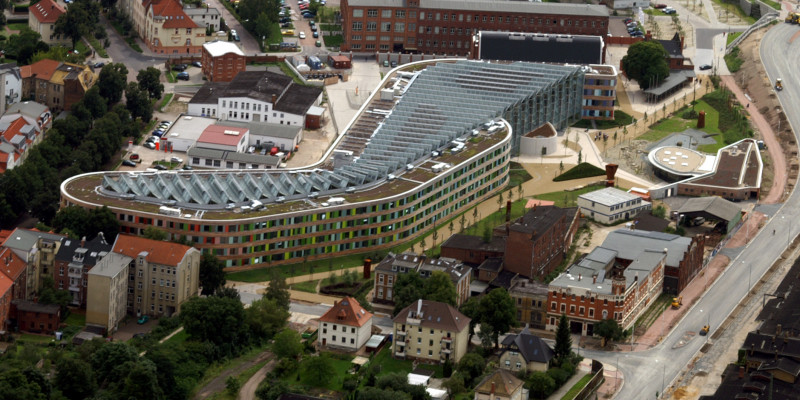 das UBA-Gebäude in Dessau-Roßlau von oben fotografiert, das Gebäude ist langgestreckt, hat eine Fassade aus Holt und bunten Glasplatten und schräggestellte Solarzellen auf dem gesamten Flachdach