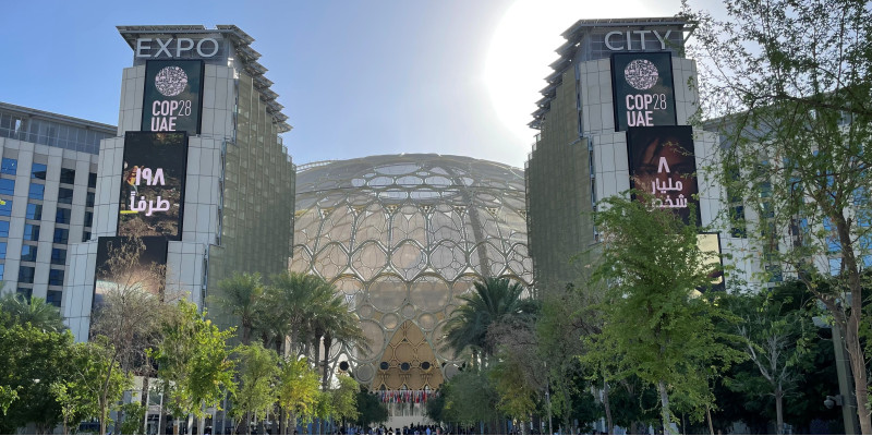 Menschen streben dem Eingang eines modernen Gebäudes mit der Aufschrift Expo City COP 28 UAE zu, auf der Straße sind Bäume in Kübeln als Allee aufgestellt
