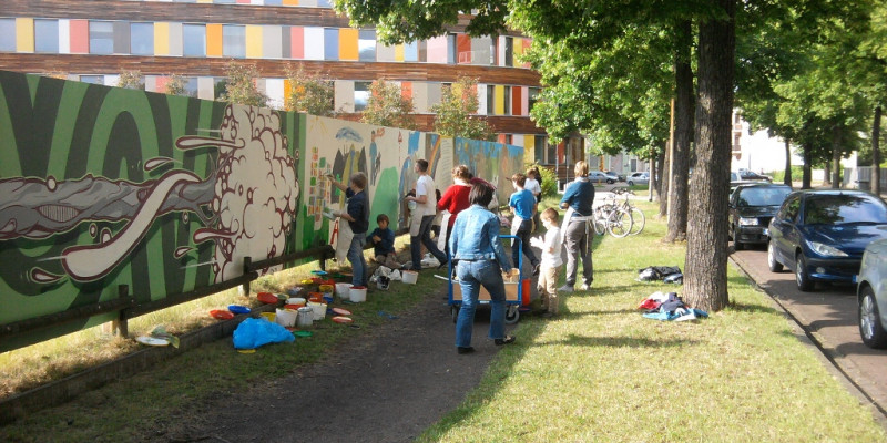 Kinder und Jugendliche malen mit Betreuerinnen den aus einer blickdichten Holzwand bestehenden Bauzaun mit bunten Bildern an