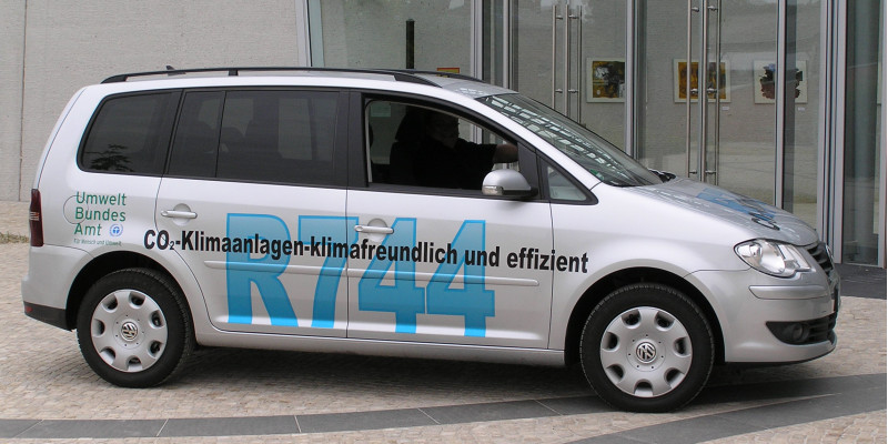 ein silbergrauer VW Touran steht vor dem UBA Dessau-Roßlau. Er trägt die Beschriftung "CO2-Klimaanlagen - klimafreundlich und effizient, R744" und das Logo des Umweltbundesamtes
