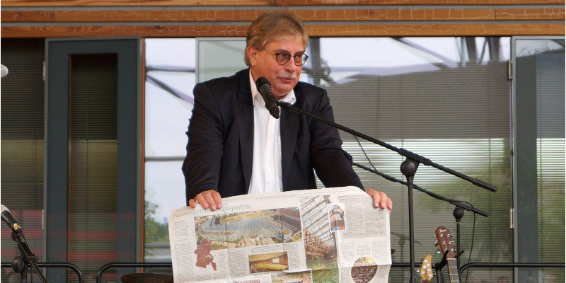 UBA-Vizepräsident Dr. Thomas Holzmann am Rednerpult. Er zeigt einen zweiseitigen Zeitungsartikel mit Fotos des UBA-Gebäudes.