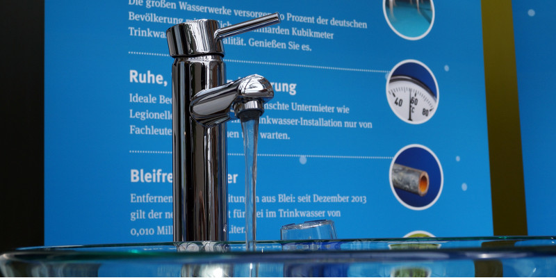 an einem Informationsstand hängt ein Plakat mit Verbrauchertipps rund um das Trinkwasser, aus einem Wasserhahn läuft Wasser in eine Glasschüssel