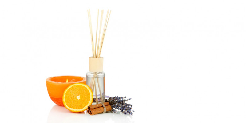 eine orange Duftkerze, ein Aromafläschchen mit Holzstäbchen zur Raumluft-Beduftung, eine halbe Orange, ein Lavendelsträußchen und Zimtstangen