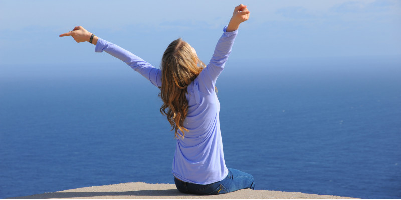 eine Frau sitzt am Meer und atmet tief durch, bis zum Horizont ist nur blaues Wasser zu sehen