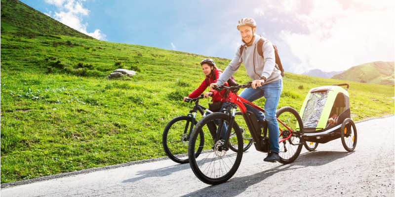 Mann auf einem E-Bike mit Kinder-Fahrradanhänger auf einer Straße in bergigem Gelände bei Sonnenschein