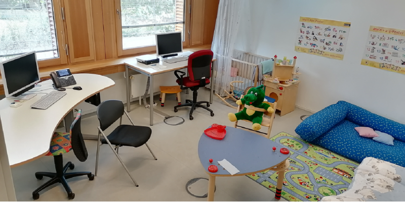 Bürozimmer mit zwei PC-Arbeitsplätzen, einem Kindertisch, Babystuhl, Sofa, Gitterbettchen, Spielteppich und Stofftier