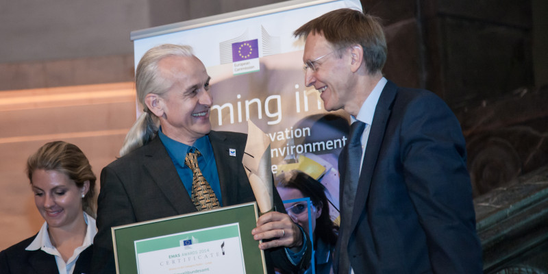 zwei lächelnde Männer vor einem Plakat mit der EU-Flagge, der eine hält ein gerahmtes Zertifikat in den Händen