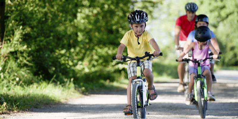 ein Mann und drei Kinder (alle mit Fahrradhelm) fahren in grüner Umgebung Fahrrad