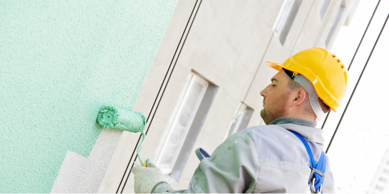 Man in overalls and yellow hard hat applying a mint green coat to a building façade