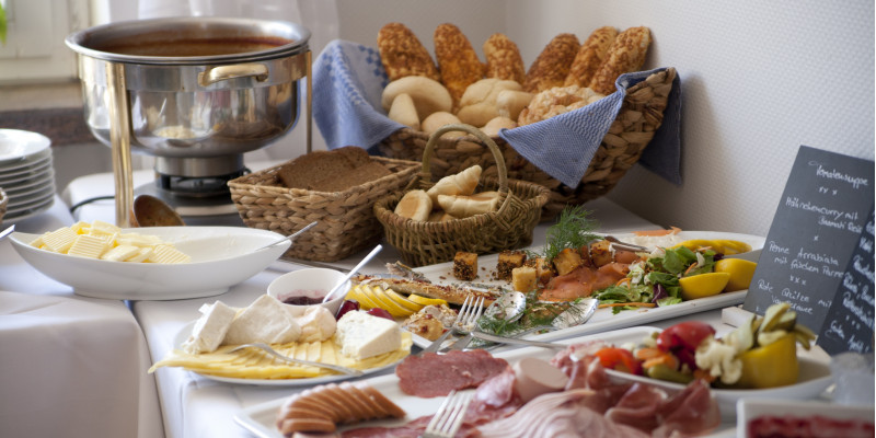 Frühstücksbuffet im Hotel mit Brötchenkorb und Porzellanplatten mit unverpackten Lebensmitteln wie Butter, Käse und Wurst-Aufschnitt