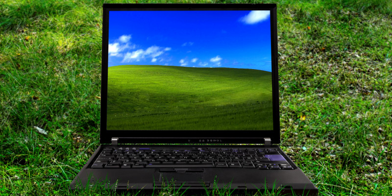 ein Laptop steht auf einer grünen Wiese, auf dem Bildschirm ist ein grüner, grasbewachsener Hügel vor blauem Himmel zu sehen