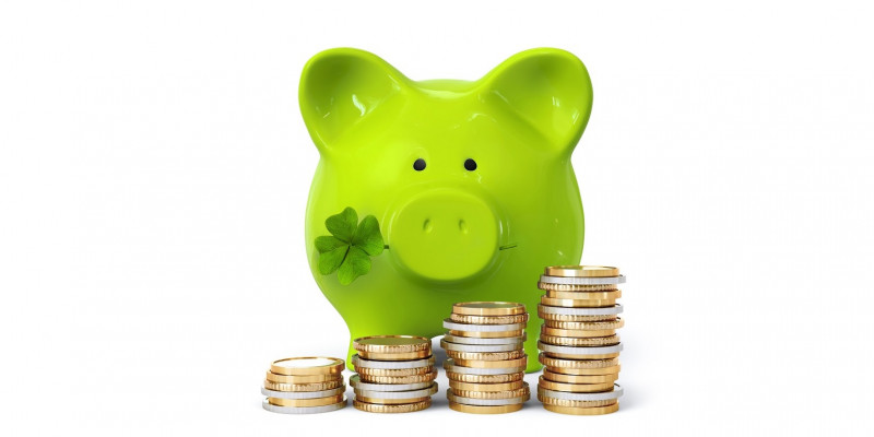 vor einem grünen Sparschwein mit einem vierblättrigen Kleeblatt im Maul sind Geldmünzen aufgestapelt