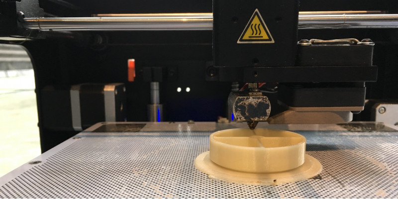 auf einer gelochten Metallplatte ist ein Plastikgegenstand zu sehen, über den der Druckkopf eines 3D-Druckers fährt