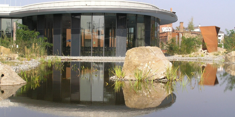 Low, round building with glass façade reflected in a pond surrounded by natural stones