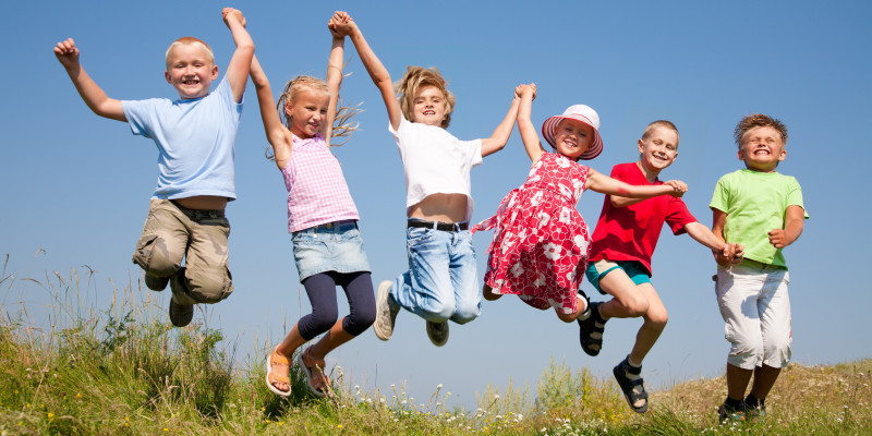 Six children on a lawn sich in einer Kette bei den Händen gefasst und hüpfen lachend in die Luft