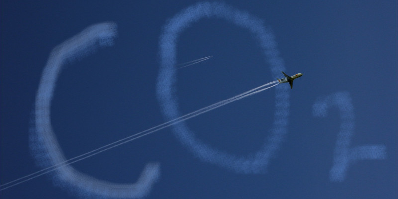 an einem blauen Himmel bilden Wolken den Schriftzug "CO2", ein Flugzeug fliegt durch das Bild und zieht einen Kondensstreifen hinter sich her