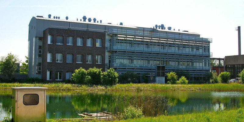Dreistöckiges, langgezogenes modernes Bürogebäude, davor ein Fluss oder Kanal mit grasbewachsenen Ufern