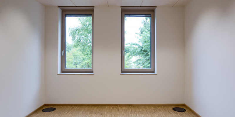 leerer Büroraum mit weißen Wänden und Holzfußboden, durch die zwei Fenster sind grüne Baumkronen zu sehen