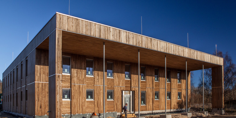 würfelförmiger, zweistöckiger Bau mit Holzfassade und Kollonadengang, davor ein Bauarbeiter, die Außenanlage noch ungestaltete Baustelle