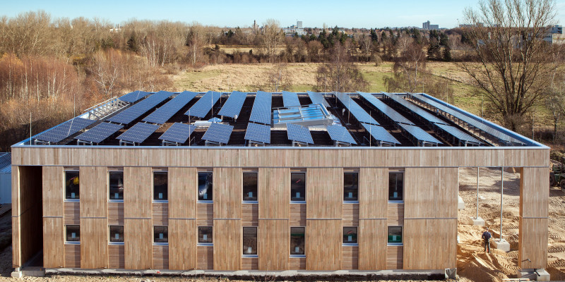 würfelförmiger, zweistöckiger Bau mit Holzfassade, auf dem gesamten Flachdach befinden sich leicht schräg aufgestellte Fotovoltaik-Elemente