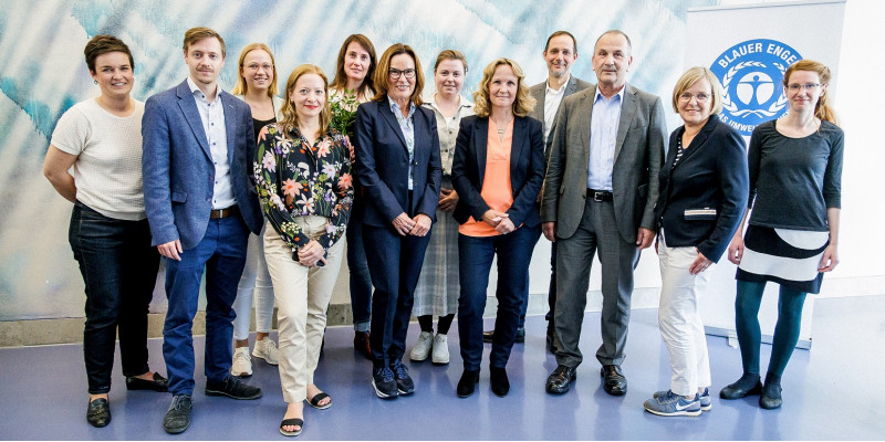 Neun Frauen und drei Männer haben sich vor einer Stellwand mit dem Logo "Blauer Engel" zu einem Gruppenfoto aufgestellt