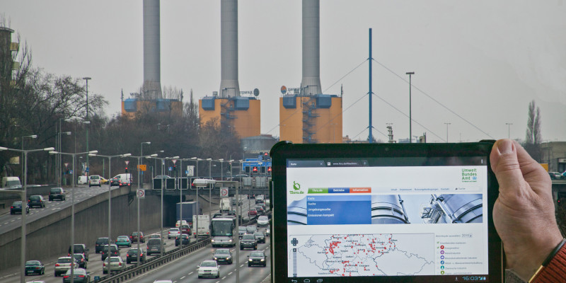 eine Hand hält ein Tablet mit dem Webangebot thru.de vor ein Kraftwerk hinter einer Stadtautobahn