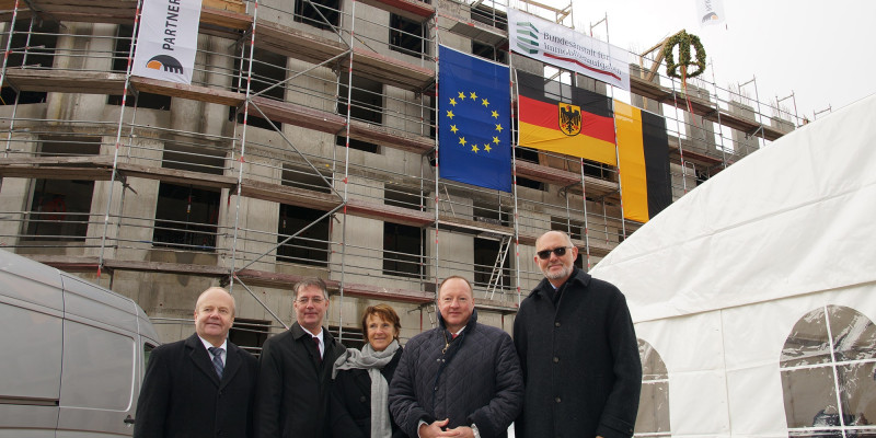 Gruppenfoto: vier Männer und eine Frau vor einem eingerüsteten Rohbau mit Richtkranz und Fahnen der EU, Deutschland, Sachsen-Anhalt, Bundesanstalt für Immobilienaufgaben und Partnerbau
