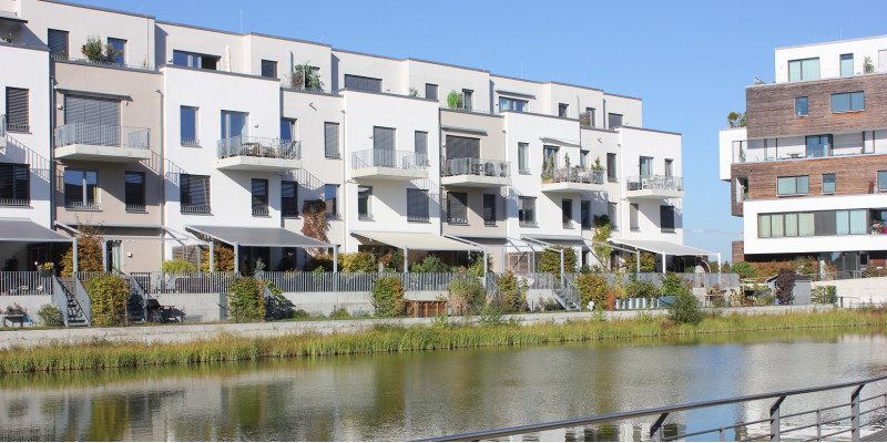 an einem Kanal in der Stadt stehen weiße, vierstöckige Häuser im Bauhausstil mit Dachterrassen, Balkonen und kleinen Gärten mit Sonnenschutz