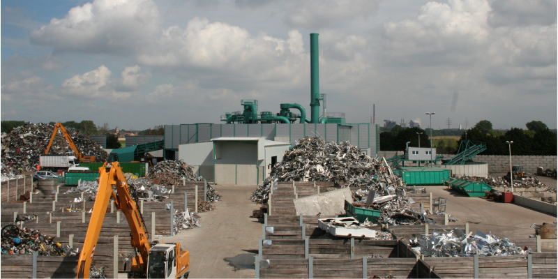Betriebsanlage mit Metallschrott, auf der Bagger arbeiten, im Hintergrund eine Schredderanlage