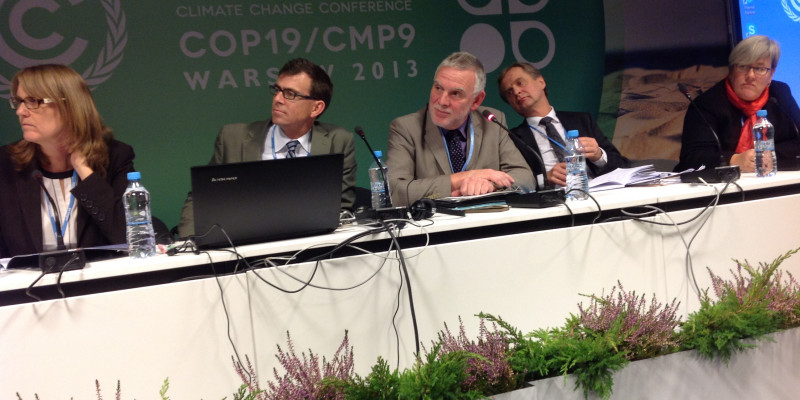 Jochen Flasbarth mit anderen Rednern auf einem Podium, im Hintergrund eine Stellwand mit dem Logo der Warschauerr Klimakonferenz COP19/CMP9