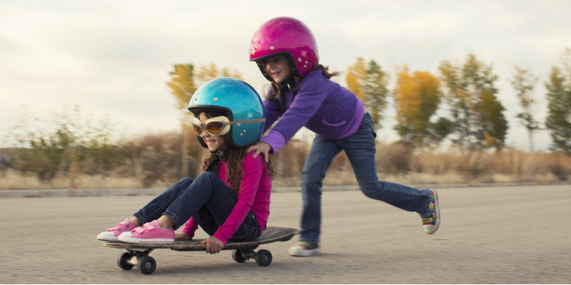 zwei kleine Mädchen mit Sturzhelmen schieben sich gegenseitig auf einem Skateboard auf einem gepflasterten Platz