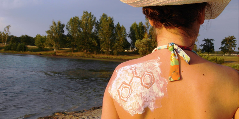 Eine Frau mit Sonnenhut und Bikini steht an einem See. Man sieht Rücken und Hinterkopf. Das linke Schulterblatt ist mit Sonnencreme eingecremt, in die eine chemische Strukturformel gezeichnet ist.