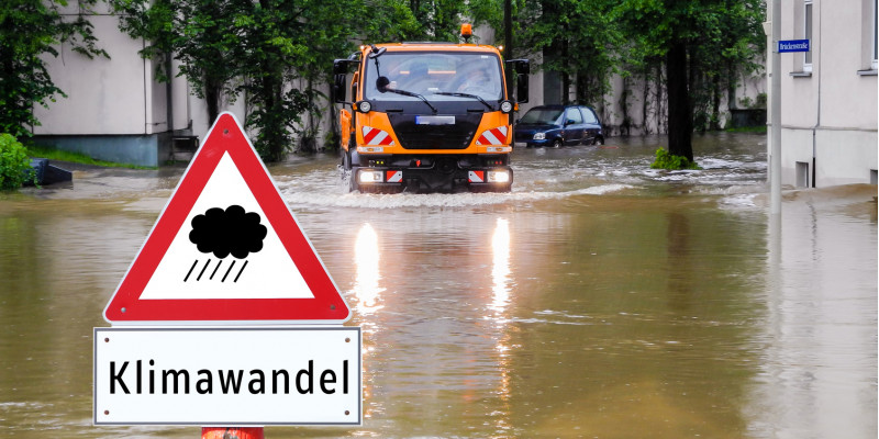 überflutete Straße in der Stadt, davor ein Warnschild "Regen / Klimawandel", ein kommunales Einsatzfahrzeug bahnt sich den Weg durchs Wasser