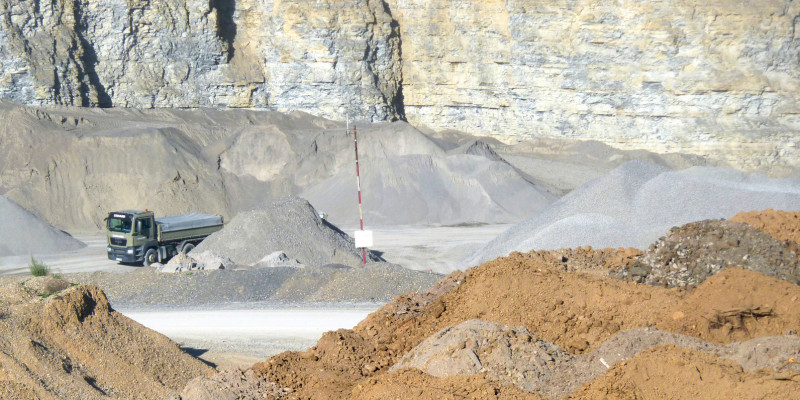 in einem Steinbruch werden Baustoffe abgebaut, man sieht verschiedenfarbige Schüttguthalden und einen Lkw vor einer steilen Felswand