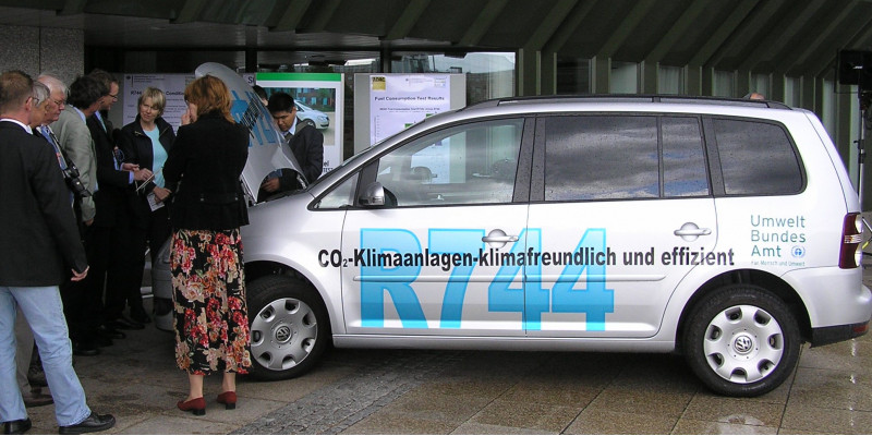 silbergraues Auto mit der Aufschrift "CO2-Klimaanlagen - klimafreundlich und effizient, R744" und dem Logo des Umweltbundesamtes, daneben eine Gruppe Menschen