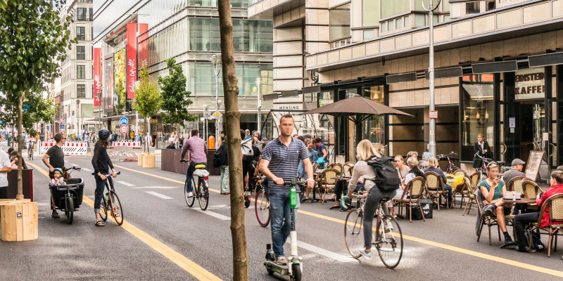 Fußgängerzone mit Radspur, Tischen vor Cafés, Geschäften und Bäumen in Kübeln mit Sitzgelegenheiten