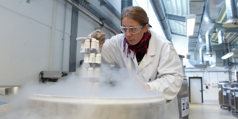 eine Frau in einem weißen Kittel, mit Schal, Handschuhen und Schutzbrille holt Proben aus einem dampfenden Metallbehälter, der in einer Halle steht