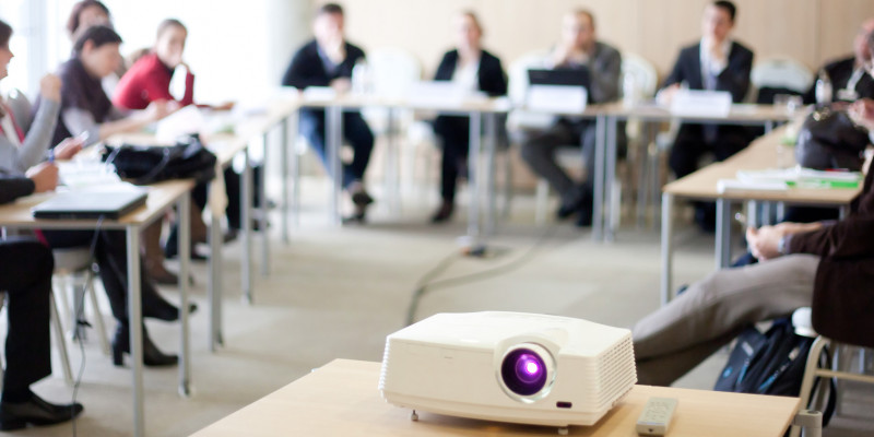 etwa 10 Leute sitzen auf einer Veranstaltung an U-förmig aufgestellten Tischen mit Namensschildern und Laptops, vorne läuft ein Beamer