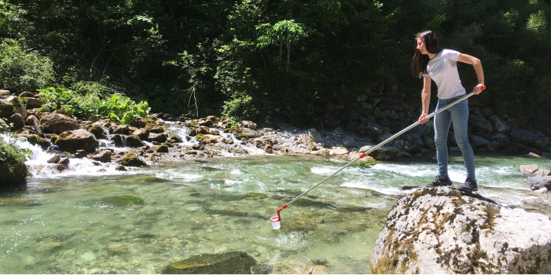 eine junge Frau steht an einem Gebirgsfluss auf einem Felsen und entnimmt mit einer Teleskopstange eine Wasserprobe aus dem Fluss