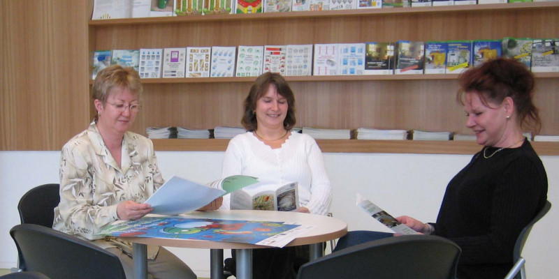 Three women sitting at a round table looking at flyers and brochures, a shelf with more materials in the background
