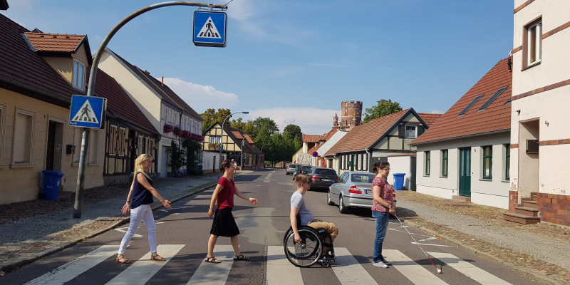 Straße mit niedrigen Fachwerkhäusern und einem Zebrastreifen, der von einer Frau mit Blindenstock, einem Rollstuhlfahrer und zwei weiteren Fußgängerinnen überquert wird.