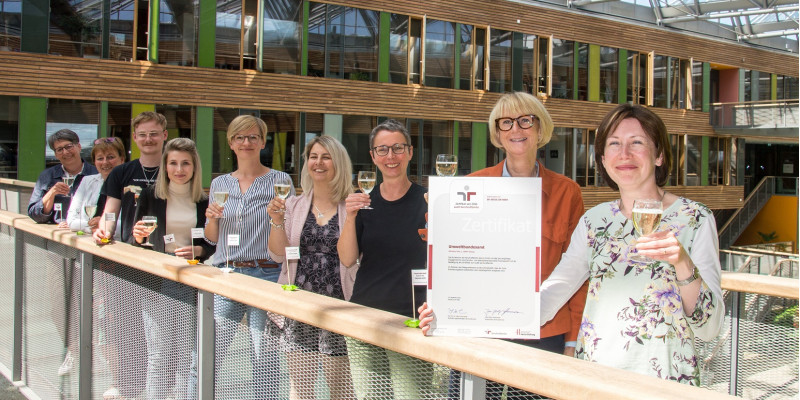 acht Frauen und ein Mann haben sich im UBA Dessau für ein Foto aufgestellt und halten lächelnd Gläser zum Anstoßen und das Zertifikat des Audits Beruf und Familie in die Kamera