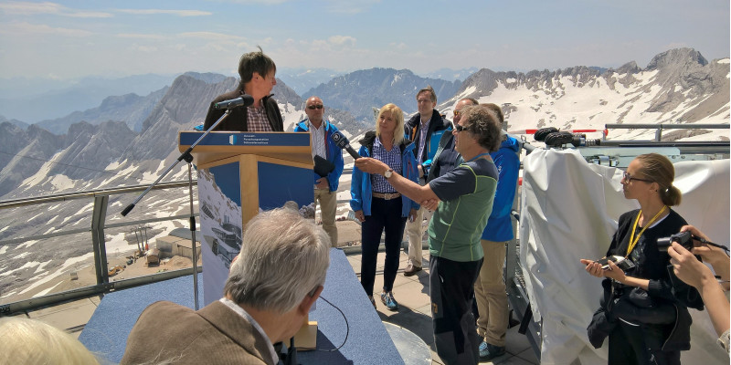 Barbara Hendricks steht auf einer Aussichtsplattform in den Bergen an einem Rednerpult mit Mikrofon, darum versammelt Reporter und andere Personen. Auf den umliegenden Gipfeln liegt Schnee.