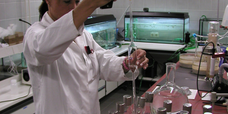 Eine Frau im Chemiekittel arbeitet im Labor. Sie befüllt Glasgefäße mit Flüssigkeit.