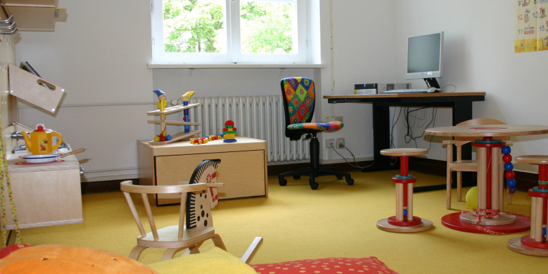 Eltern-Kind-Zimmer: Am Fenster steht ein Schreibtisch umgeben von Spielsachen, einer Spielküche, einer Sitzecke, im Vordergrund eine Matratze mit großem Kissen.